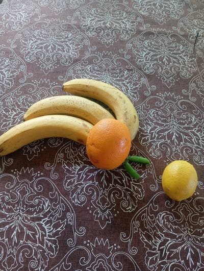 The image displays a still life arrangement of fruits and vegetables on a brown tablecloth with a white ornate pattern. The subjects are positioned in the lower half of the frame. Three yellow bananas are clustered together, curving from the left towards the center. To the right of the bananas sits a bright orange. Below the orange, two small green pea pods are visible. Further to the right, a single yellow lemon rests on the tablecloth. The lighting appears to be natural, casting soft shadows. The scene is static, with no discernible action or interaction. The location in Pivdennoukrainsk, Ukraine, cannot be determined from the visual information provided in the image.