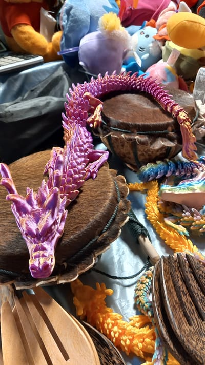 A purple and gold articulated dragon figurine rests on a brown, fur-covered drum. Another articulated dragon, in shades of blue, green, yellow, and purple, is partially visible in the background. A bright orange articulated dragon is also present. In the foreground, a wooden salad fork and tongs are laid out on a light blue, textured surface. Several stuffed animals, including Winnie the Pooh and a blue character with yellow accents, are scattered in the background. The scene appears to be an indoor setting.
