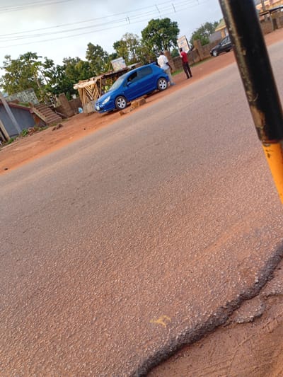 The image depicts a roadside scene in Kuru Dungus, Nigeria, with a clear view of a blue car parked on the side of a dusty, reddish-brown road. Two men are visible near the car. One man, wearing a white shirt and dark pants, stands beside the blue car, while another man in a red shirt and dark pants stands further down the road, facing away from the camera. In the background, there is a small structure with a thatched roof, possibly a vendor stall, and dense green foliage. Another dark-colored car is parked on the opposite side of the road, further in the background. The sky is overcast. The foreground of the image is dominated by the textured surface of the road.