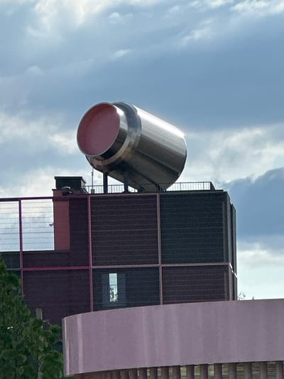 This image captures a large, prominent industrial-style cylindrical structure mounted atop a building in Бараўлянскі сельскі Савет, Belarus. The main subject is a substantial, horizontally oriented cylinder crafted from a reflective, brushed metallic material, likely stainless steel or aluminum. Its open end, positioned towards the upper-left, reveals a smooth, matte pink interior. The cylinder is supported by a dark, open metal framework that includes railings, situated on the rooftop of a multi-story building.
The building itself features dark gray or brown facades with a textured surface, possibly composed of brickwork or panels. These dark sections are segmented by reddish-brown vertical and horizontal beams or decorative elements. On the rooftop, directly beneath the large cylinder, a smaller, dark green rectangular unit is visible, alongside metal railings. To the left side of the building, a fine mesh fence partially obscures a white, oval-shaped object mounted on a pole, possibly a surveillance camera or sensor.
In the lower foreground, a smooth, curved structure in a light pink or lavender tone occupies a significant portion of the frame. Below this, a dark railing or fence is partially visible. To the far left, green foliage from a tree or bush is present. The background consists of a bright blue sky interspersed with scattered white and light gray clouds, indicating daylight conditions.