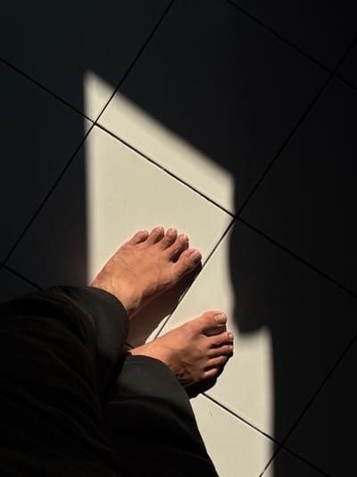 The image shows two bare feet positioned on a tiled floor, with a sharp diagonal shadow cast across them. The tiles appear to be light-colored and square, arranged in a grid pattern. One foot is closer to the viewer, with toes clearly visible, and is partially covered by dark fabric, likely pants. The other foot is further away and mostly illuminated by sunlight. The lighting suggests a strong light source, possibly from a window or an opening, creating a high contrast between the illuminated and shadowed areas of the floor. There are no other people or objects visible in the frame. The scene is likely indoors or in a covered outdoor space due to the tiled floor and the nature of the shadow. The location is specified as Kalirancang, Indonesia.