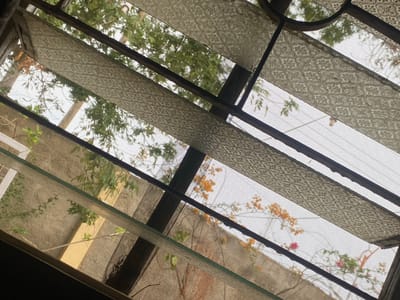 The image depicts a view through a louvre window in Giginyu, Nigeria. The window features patterned glass panes and mesh screens, obscuring the details of the exterior. Outside, greenery from trees is visible, along with a beige wall and a plant with orange flowers. The lighting suggests it is daytime, with sunlight filtering through the foliage. No people or specific activities are discernible. The scene appears to be a residential or domestic setting, typical of the Giginyu area.