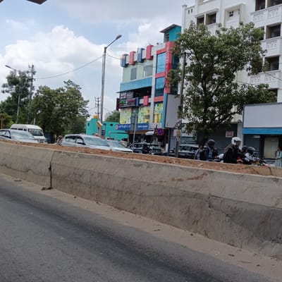 This image captures a bustling street scene in Bengaluru, India, under a cloudy daytime sky. The foreground features a concrete road divider or barrier, constructed with rough concrete blocks, separating the viewer's perspective from a service road or parking area. On the service road, several parked or slowly moving vehicles are visible, including multiple cars (one silver SUV-like car prominent on the right), a white van further back, and several motorcycles.

People are present in various parts of the scene. On the right side of the service road, several individuals are on motorcycles, wearing helmets. One person in a white helmet is prominently positioned on a motorcycle, looking forward. Further to the left, a person is standing by a white car, and another person with what appears to be a head covering is visible in the distance near a white van. A woman in a light-colored dress is standing near the right edge of the frame.

The background is dominated by commercial buildings and lush green trees. A multi-story commercial building with colorful facades (red, blue, light blue, and grey panels) stands out. Visible signs on this building include "SRI SAI PLAZA" on its side, "NINAN'S STEAKHOUSE," "NEVER SPA," and "HDFC Bank." Below HDFC Bank, there is text in Kannada script. Another, larger white building with a more traditional architectural style is seen to the right, partially obscured by a large tree. A sign on this white building indicates "COMMERCIAL SPACE FOR LEASE." Utility poles with numerous wires and streetlights are scattered throughout the image. A red and blue "No Stopping" or "No Parking" sign is also visible. The sky is overcast with white and grey clouds.