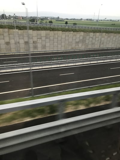 The image depicts a multi-lane highway segment, observed from an elevated perspective, likely from an adjacent overpass or parallel road, near Adjud, Romania. The foreground features a blurred silver metal guardrail, below which is dark, indistinct ground. Immediately beyond, a strip of green grass shows motion blur. The main subject is a newly paved black asphalt highway, comprising at least two lanes in each direction, separated by a median strip. The lanes are marked with crisp white solid and dashed lines. A silver metal guardrail runs along the right edge of the highway, separating it from the median. Within the median, a series of vertical green posts are regularly spaced, possibly supporting a flexible barrier or signage.
To the left of the highway, a substantial retaining wall constructed from large, rectangular concrete blocks, varying in shade from light to medium grey, supports an elevated terrain. Above this retaining wall, a patch of green vegetation is visible, followed by a level area featuring a silver metal barrier fence. Beyond this fence, the landscape transitions into broad green fields interspersed with scattered rural residential structures. In the distant background, a low range of hills or mountains stretches horizontally under an overcast sky. Multiple utility poles or streetlights are sparsely distributed along the highway's side, some exhibiting motion blur. No people, vehicles, or specific interactions are discernible within the frame.