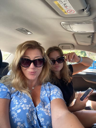 The image shows two individuals inside a vehicle, likely a car, during the daytime. The environment appears to be a sunny day, with greenery visible through the car windows. The individuals are positioned in the back seat. The person in the foreground is a woman with blonde hair, wearing sunglasses and a blue floral dress. She is looking at the camera and appears to be making a facial expression. Beside her, a younger person with blonde hair, also wearing sunglasses, is looking towards the camera and holding one hand up near their sunglasses, possibly adjusting them. This person is wearing a black t-shirt. A smartphone is visible in the hands of the person on the right. The interior of the car shows a sun visor with an airbag warning label and a light fixture on the ceiling. The presence of trees and some structures outside suggests a location that is not densely urbanized, consistent with the description of Palje Brdo, Croatia, which is a rural area. The image is a selfie-style shot, capturing a moment of travel or outing.