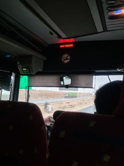 The image captures an interior view from a vehicle, likely a bus or coach, traveling through the city of Al-Kiswa, Syria. In the upper portion, a red LED display panel reads "ARRET DEMANDE" (Stop Requested) in French, positioned above a circular "No Smoking" pictogram. A device with a green-lit screen is mounted to the upper left of the windshield area. Two red, patterned seats are partially visible in the immediate foreground, with a dark headrest on the right seat. Beyond the windshield, a wet, two-lane road stretches through a barren, dry landscape. A green truck is visible in the middle distance on the road, followed by a lighter-colored vehicle further ahead. A blurred figure is present on the dirt shoulder to the left of the road. The sky appears overcast.