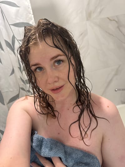 A woman with wet dark hair and blue eyes is partially visible in what appears to be a bathroom. She is holding a gray towel close to her body. The background includes a marble-like wall and a shower curtain with a leaf pattern. The location is Ферма, Russia.