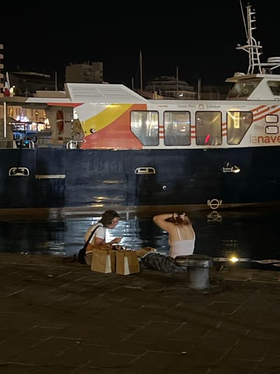 In Marseille, France, at night, two individuals are seated on a stone quay next to a large ferry. The ferry, displaying the colors of the French flag on its side, is docked in dark water. The first person, on the left, is wearing a white t-shirt and dark pants, with a black bag slung over their shoulder. They are looking down at a mobile phone and a paper bag. The second person, on the right, is wearing a light-colored tank top and dark pants. They are seen from behind, with their hands in their hair. In front of them are two brown paper bags. A large metal cleat is positioned between them and the water. The quay is paved with large, dark stones, some of which are wet and reflect the ambient light. The background shows buildings and other vessels, dimly lit against the dark night sky.