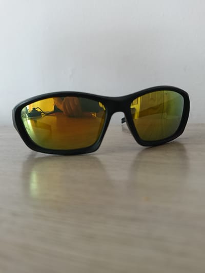 A pair of black-framed sunglasses with yellow mirrored lenses rests on a wooden surface. The lenses reflect a hand and a cord with a black object, likely a cable or charger, on a white wall. The surface appears to be a table or desk. The lighting is soft, with a shadow cast by the sunglasses. The background is a plain white wall. The location is not discernible from the image.