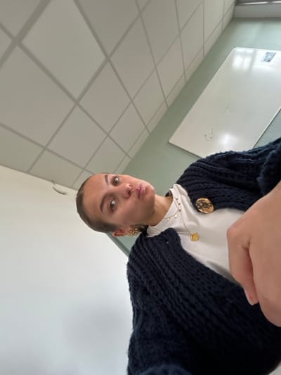 The image presents a close-up, low-angle, and slightly tilted portrait of a young adult female indoors. She has brown eyes, light brown hair pulled back, and is making a 'duck face' expression while looking directly at the camera. She wears a white t-shirt, a dark navy or black chunky knit cardigan secured by two large, circular gold-toned buttons or brooches on the right shoulder area, multiple gold-toned necklaces (including one with a flat circular pendant and others with small disc embellishments), and a visible gold-toned earring on her left ear.
The background consists of a white drop ceiling with square acoustic panels set within a light grey grid. The wall to her left is plain white, contrasting with the wall to her right, which is painted a light teal or mint green. A large, rectangular white dry-erase board is mounted on the green wall, bearing faint blue markings, including the alphanumeric "S23," a simple drawn smiley face, and other indistinct symbols. The environment is consistent with an institutional setting such as a classroom, conference room, or office. No specific landmarks or identifying features are present in the image to pinpoint the exact location within the city of Saint-Étienne, France.
