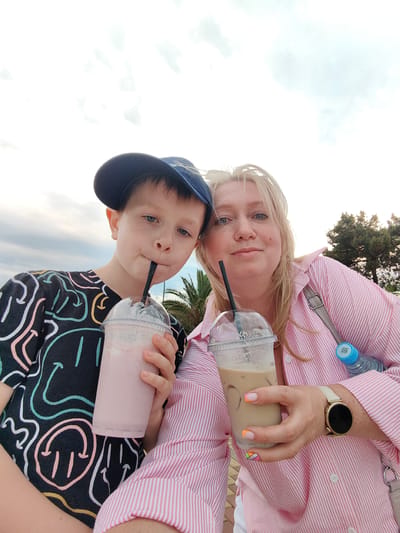 A boy wearing a blue baseball cap and a black t-shirt with a colorful smiley face pattern holds a pink milkshake. Beside him, a woman wearing a pink and white striped button-up shirt holds a coffee drink. They are both looking at the camera, smiling. The background shows a cloudy sky and some trees. The location is in the city of Sirius, Russia.