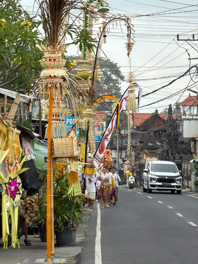 The image shows a street scene in Mengwi, Indonesia, with a procession or ceremony taking place. A group of people, mostly women, dressed in traditional Balinese attire, are walking along the right side of the road. Some are carrying decorative items, possibly offerings. There are also several tall, ornate, woven structures with palm fronds and colorful decorations lining the street, suggesting a festive or religious event. A white SUV is visible on the road, as well as a scooter. The buildings in the background appear to be traditional Balinese architecture with tiled roofs. Overhead, a dense network of electrical wires crisscrosses the sky. The overall atmosphere is one of cultural activity within an urban environment.