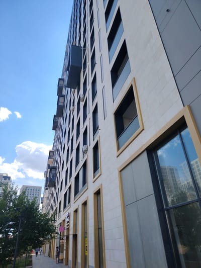 This is a wide-angle, eye-level shot of a modern apartment building on a bright, sunny day. The building dominates the frame, its facade a mix of light stone tiles and dark gray panels. Numerous windows are visible, many of them recessed and framed with gold trim, creating a geometric pattern. Some windows have dark balconies protruding outwards. Air conditioning units are mounted on the exterior walls between windows. A large, dark-framed window on the ground floor reflects the blue sky and clouds, as well as other buildings.

To the left of the building, a sidewalk runs alongside a small green space with trees and a lamppost. In the distance, other apartment buildings can be seen. Several people are walking on the sidewalk, some dressed casually. The atmosphere appears relaxed and everyday. A few signs are visible on the building's lower levels, including one with Cyrillic text in pink and white, and another yellow sign with text. The time of day appears to be midday, given the strong sunlight and clear blue sky with scattered white clouds. The location is implicitly Astana, Kazakhstan, as indicated in the prompt.