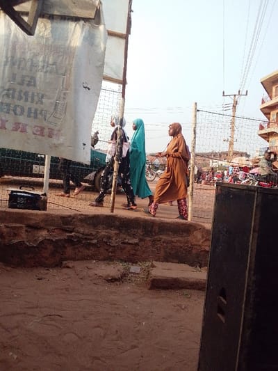 Three Muslim women walk along a raised pathway. One wears a white hijab and patterned pants, another wears a turquoise hijab and dress, and the third wears a brown hijab and dress with patterned pants. Behind them is a chain-link fence, street traffic, and a building in the distance. The pathway has a short, crumbling wall, and the ground is dirt. A large sign and a speaker are visible on the left and right of the frame.