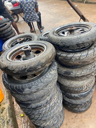 The image displays a collection of used motorcycle tires, some mounted on rims, stacked in an outdoor setting. The tires are black and appear worn, with visible tread patterns. Several individuals are partially visible in the frame, suggesting their involvement in handling or organizing the tires. One person, wearing a blue and white patterned shirt and black pants, is seen from the back, sitting on what appears to be a small cart or vehicle. Another person's arm is visible, reaching towards the tires. The ground is a dirt or concrete surface, wet in places, indicating a possible recent rain or cleaning activity. A small green object, possibly a lime or small fruit, rests on the wet ground near the tires. A metal stand or rack is present in the background, supporting some of the tires. The overall impression is of a tire storage or sale area, likely in a workshop or marketplace. The lighting suggests it is daytime, but there are no strong shadows indicating a specific time. The presence of multiple tires and the apparent activity suggest a commercial or repair-related context. The location is specified as Ogidi, Nigeria, which provides a cultural and geographical context for the scene. There is no discernible text in the image.