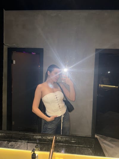 A young woman is seen in a mirror selfie. She is wearing a white corset top and jeans. The background includes a concrete wall, black doorframes, and a glimpse of a metallic fixture. The location appears to be an indoor space, potentially in Odintsovo, Russia, but details are insufficient to confirm the exact location. The subject is taking a photograph of herself using a mobile phone.
