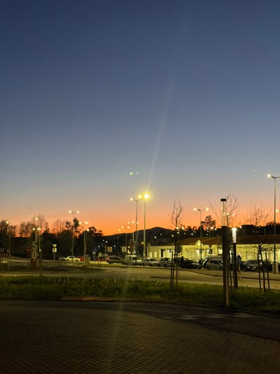 An outdoor scene in Penafiel, Portugal, is depicted at dusk or sunset, characterized by a sky gradient transitioning from deep blue overhead to a vibrant orange-red near the horizon. The foreground consists of a dark, paved surface, likely a road or parking lot entrance, separated by a narrow strip of dark green grass and several young, bare-branched trees.
In the midground, a multi-lane road segment or intersection is visible on the left, featuring a tall brown signpost, a white circular traffic sign with a red border and diagonal line, and a "50" speed limit sign. Beyond this, a dense line of trees with green foliage ascends a gentle slope. To the right, a parking lot contains approximately twenty parked passenger vehicles of various makes and colors. Adjacent to the parking lot are several single-story buildings with light-colored facades and pitched reddish-brown roofs, indicative of commercial or administrative use. One building prominently displays "ANEL NOGUEIRA" on its facade.
The scene is illuminated by numerous tall streetlights emitting a bright yellow glow, casting light across the parking area and buildings. A prominent lens flare originates from the lower right, extending diagonally upwards, accompanied by two smaller, distinct green circular flares. The background reveals distant hills or mountains silhouetted against the vivid orange sky, sparsely covered with trees. No individuals or active interactions are observed within the frame; the scene is static.