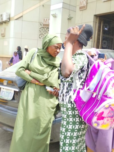 The scene depicts an outdoor urban environment in Sabon Gari East, Kano State, Nigeria, during daylight hours. The primary focus is on three individuals in the foreground. On the left, a woman in a light green hijab and matching long-sleeved dress holds a white smartphone in her right hand, her left arm crossed over her midsection; she faces towards the right with a serious expression. Adjacent to her, a second woman, partially visible from the chest up, wears a black knit beanie and a white and dark green patterned dress, with her right hand raised and fingers curled as if in mid-gesture or speech. Behind this second woman, a third individual, partially obscured, wears a light purple hijab and holds a dark-colored smartphone, directed towards the scene. A bright pink backpack with multi-colored floral designs is prominently displayed in the lower right foreground. Behind the woman in green, a dark-colored sedan is partially visible, displaying a Nigerian license plate: "KANO PCH224TV." The background features a light-colored building with windows and air conditioning units, bearing multiple "MD MUDATEX" signs. Further in the distant left background, additional individuals, including children and a man wearing a light hat, are visible.