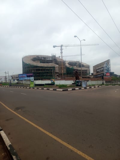 This image depicts a large, unfinished building under construction in Enugu, Nigeria. The building has a distinctive curved, spiral design and is surrounded by scaffolding. Two large cranes are visible, indicating ongoing construction work. In the foreground, a road with yellow lane markings runs past the construction site.
Several billboards are positioned along the road. One prominent billboard advertises an "ACCELERATE CONFERENCE" with the subtitle "PASTORS & LEADERS RETREAT." Another billboard features an artist's rendition of a modern building complex with the text "Own a piece of the Enugu Skyline." A smaller green billboard states "THE SKY'S NO... ENUGU STATE." A statue of a person is also visible near the construction site.
The weather appears overcast, suggesting a cloudy or possibly rainy day. The time of day is not explicitly indicated, but the general lighting suggests it could be daytime. The scene is urban, with the road and billboards indicating a public space in a city. The overall impression is one of development and progress, with a focus on a major construction project. The image captures a moment in the transformation of the urban landscape of Enugu.