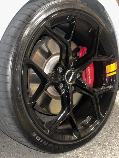 A close-up, oblique view focuses on the front passenger side wheel and tire assembly of a white vehicle, likely a Toyota Supra given the visible "SUPRA" branding on the brake caliper. The wheel is a multi-spoke design with a glossy black finish, secured by five visible lug nuts. A "SIGMA" branded center cap is present. Behind the spokes, a large, silver-toned metallic brake rotor and a prominent red brake caliper are visible. The caliper also features an orange and yellow two-segment stripe on its right side. The tire is a black rubber "GOODRIDE SUPERCUP" model with the word "GOODRIDE" visible on the sidewall, and appears to show moderate tread wear. The tire's valve stem is present in the lower right quadrant of the wheel. The vehicle is positioned on a dark, textured asphalt or paved surface. No individuals, actions, or specific interactions are depicted. The lighting suggests an indoor or sheltered outdoor environment. The specific location within Volketswil, Switzerland, cannot be determined from the visual information provided.