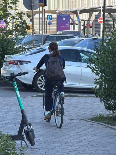 In Astana, Kazakhstan, a person is riding a bicycle on a paved walkway. The cyclist, viewed from behind, is wearing a dark jacket, dark pants, and socks with green accents. A dark backpack is worn on their back. The bicycle has a blue frame and is equipped with a rear fender. In the foreground, a green electric scooter with "Bolt" branding stands upright. Behind the cyclist, a silver sedan is parked. The background features a modern building with a ribbed facade, an overpass structure, and traffic signs. Several other cars are visible in traffic lanes. Green foliage from trees and bushes is present on the sides of the walkway and near the building. The scene takes place during daylight.