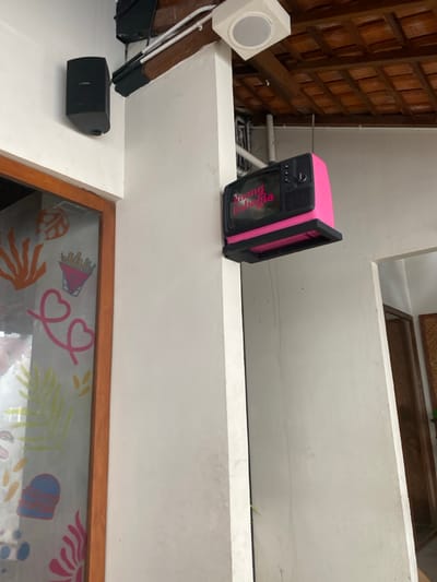 The image shows the exterior of a building in Surakarta, Indonesia, located at the coordinates -7.557 latitude and 110.796 longitude. A pink, faux vintage television, mounted to a white wall, with the words "Ruang Bahagia" (which means "Happy Room") written on it, is the focal point. The TV is mounted to a wall, which is part of a corner of the building. Another wall meets the corner, and an open doorway with a wooden frame leads into a space with a white wall. To the left of the television, a wooden door frame, leading into another space, has a glass panel that displays colorful artwork on it. In the background, a ceiling with a tiled, brown surface is visible above the doorway and the television. A white ceiling speaker can be seen in the upper left of the image. There are no people present in the image.