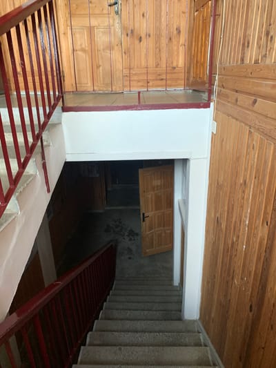 The image depicts the interior of a stairwell in Kapyĺ, Belarus. Red metal railings line a concrete staircase descending into a dimly lit area. To the right, a wall is covered in vertical wooden planks. A white wall extends across the top of the stairwell opening. Further down the stairs, a wooden door is ajar, revealing a dark interior. The overall impression is that of a functional, somewhat aged, building interior.