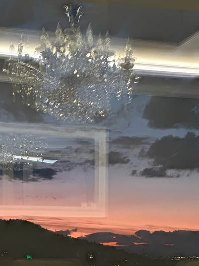 The image depicts a reflection of a crystal chandelier superimposed over a sunset scene viewed through a window, likely in Asenovgrad, Bulgaria. The chandelier, intricately detailed with multiple tiers of crystals and metallic fixtures, is the primary subject in the foreground, occupying the upper portion of the frame. Its reflection is clear, catching the light and creating a dazzling effect.
Beneath the chandelier's reflection, the scene transitions to a landscape visible through the window. A dark, silhouetted hillside dominates the lower part of the image, suggesting a mountainous or elevated terrain. Above the hillside, the sky is filled with dramatic cloud formations and a vibrant sunset. The sky transitions from a soft pink and orange hue near the horizon to shades of purple and gray at higher altitudes, indicating the presence of both illuminated and shadowed clouds. The horizon line is marked by the dark silhouette of rolling hills and distant lights that suggest a populated area, possibly the city of Asenovgrad. The reflection of the chandelier also appears faintly in the lower part of the image, interspersed with the sunset scenery. No people are visible in the scene.