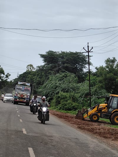 In Malkapur, India, a two-lane asphalt road is visible under an overcast sky. On the left side of the road, a white car, a decorative truck, and two motorcycles are traveling in the same direction. The truck is brightly painted with various images and text, including "GOODS CARRIER." The first motorcycle is ridden by two individuals, while the second motorcycle, closer to the camera, carries a single rider wearing a white turban. On the right side of the road, a yellow JCB excavator is parked on a dirt embankment beside the road, which appears to be undergoing some construction or repair. Dense green foliage lines the sides of the road, with trees and bushes forming a thick natural barrier. Power lines are strung across the upper portion of the image, supported by a utility pole.