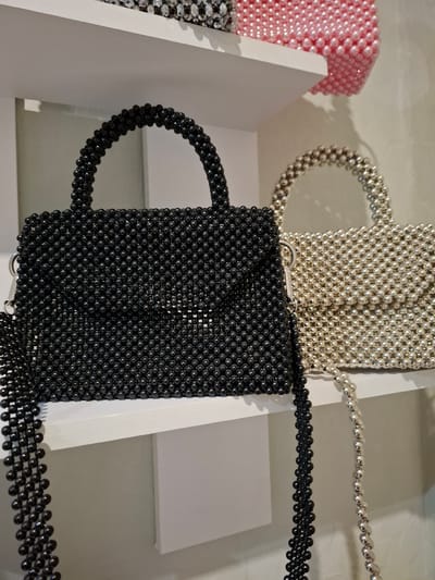 The image displays two beaded handbags on a white shelf. The handbag in the foreground is black and features a top handle and a detachable strap made of black beads. To its right, a silver beaded handbag is partially visible, also with a top handle and strap. A pink beaded handbag is visible on a shelf above. The background is a plain wall. The image context suggests this scene is located in Korenovsk, Russia.