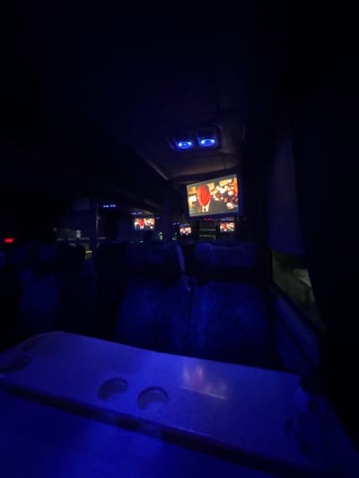The image shows the interior of a dimly lit bus, likely at night. The dominant color is a deep blue, cast by overhead lighting and possibly the screens on the bus. In the foreground, a tray table is visible, illuminated by the blue light. In the mid-ground, rows of dark bus seats are apparent, with some featuring patterned upholstery. Mounted on the ceiling of the bus, several flat-screen televisions are displaying content. One prominent screen shows a close-up of a man in a suit and tie, with a Christmas tree visible in the background. Other screens further back also appear to be displaying similar content. The environment suggests a public transportation setting, possibly a long-distance bus or a tour bus. There is no visible indication of the city of Cuernavaca, Mexico within the image itself.