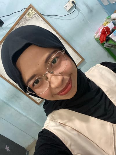 The image shows a close-up, selfie-style shot of a young woman wearing a black hijab and glasses. She is smiling slightly and looking at the camera. She is wearing a black top and a light-colored vest. Behind her, a light blue wall is visible. In the upper left corner, a framed whiteboard with some writing on it can be seen. To the right, a shelf with various items, including some plastic bags and a red object, is partially visible. The overall impression is of an indoor setting, possibly a home or office. There is no indication that the image was taken in Tanjung Gading, Indonesia.