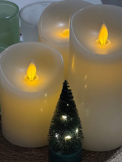 A top-down view shows three lit, flameless candles stacked vertically. The flickering lights inside are yellow. A small, artificial Christmas tree with embedded fairy lights lies horizontally across the top and middle candles. The base of the tree is a textured, dark green. The candles rest on a surface that appears to be a woven mat. The bottom of the image has glimpses of a light green glass container.