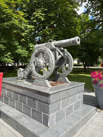 This media file shows a close-up, rotating view of a historical cannon displayed on a granite monument. The cannon, made of weathered gray metal, is ornately decorated with intricate carvings on its wheels and carriage. The monument itself is constructed from polished gray granite tiles, with a tiered design that includes steps leading up to the cannon's display base.
In the background, lush green trees provide a natural backdrop, and a patch of blue sky with white clouds is visible through the foliage. A large stone planter filled with vibrant pink flowers sits on the right side of the monument, adding a splash of color to the scene. The overall setting appears to be an outdoor public space, likely a park or memorial area, given the manicured lawn and trees.
The lighting suggests it is daytime, with clear skies and bright sunlight casting shadows, indicating fair weather. There is no visible text in the image. The media file captures a static display, suggesting no particular activity or event is taking place other than the presentation of the monument and cannon. The focus is on the historical artifact and its elaborate presentation.