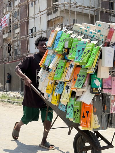 In Igboefon, Nigeria, an adult male street vendor pushes a single-wheel metal wheelbarrow laden with electronics merchandise forward on an unpaved road under bright sunlight.
The vendor, with dark skin and short, dark, unkempt hair, wears a dark t-shirt, dark green shorts, and olive-green slide sandals. He grips the wheelbarrow's handles with both hands, his gaze directed forward. The wheelbarrow features an attached metal display rack showcasing a dense arrangement of products. Merchandise includes numerous packaged wired earphones (in colorful boxes, primarily green, blue, yellow, and orange, with brands such as "DEEPER BASS," "JINDARX," "PROMAC," and "FRESH"), various white power adapters/chargers (some labeled "FAST CHARGER" or "9A"), and an assortment of plastic phone cases (clear, dark, and some pink).
In the mid-ground to the left, a second male figure stands near a light-colored concrete wall, appearing to look down at a mobile device. The background features a multi-story building under construction or renovation, enveloped by extensive bamboo scaffolding. A red banner with partially visible white text ("ONG") hangs from the scaffolding. The ground is a light-colored dirt or gravel surface, marked by shadows from the wheelbarrow and vendor.