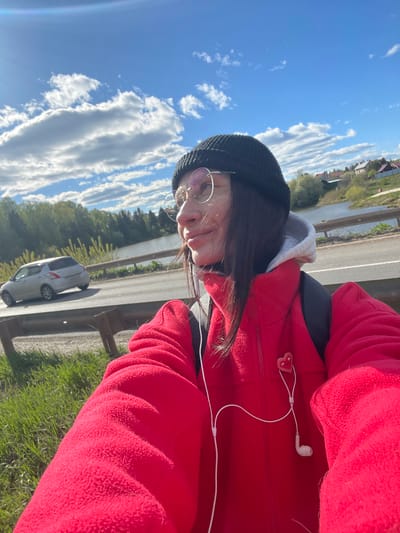 A person is seen in a medium, outdoor shot taken in Chemoshur, Russia. The individual is wearing a red fleece jacket, a black beanie, and glasses. They are positioned next to a road with a metallic guardrail. A silver car is visible on the road. In the background, there is a body of water and some vegetation under a blue sky with fluffy white clouds. The person appears to be wearing earphones, one of which is visible.