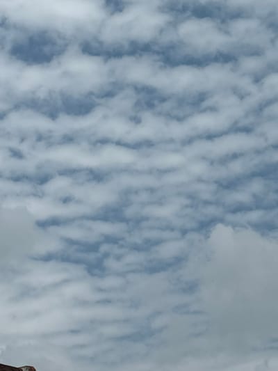 The image captures a sky filled with elongated, striated clouds. They appear like parallel brushstrokes against a pale blue canvas. The clouds are primarily white and light gray, creating a sense of texture and movement. A small section in the lower right corner seems to show the edge of a physical structure, partially obscuring the otherwise uninterrupted view of the sky.