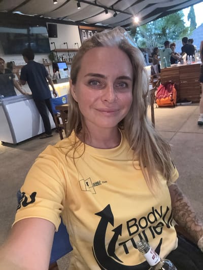 A woman with blonde hair is taking a selfie in an outdoor seating area with a bar in the background. She is wearing a yellow t-shirt with black graphics. In the background, there are several people, some of whom appear to be bar staff. There is also a television screen on the wall and a bar counter with bottles on display. The lighting suggests it is evening. The location appears to be a bar or restaurant in a tropical setting, possibly in Ban Bang Rak, Thailand.