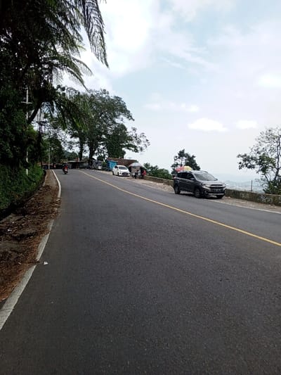 The image presents a daytime scene of a mountain road in Ciloto, Indonesia, viewed from a low angle looking forward along the road. The asphalt road occupies the majority of the frame, featuring a solid white line defining the left shoulder and a double solid yellow line marking the center. The road surface appears generally well-maintained.
On the left side of the road, a steep embankment covered in dense green foliage and trees rises. Overhanging fern-like leaves are prominent in the upper left corner. A power pole or utility structure with white components is partially visible amidst the foliage. Dirt and dry leaves are present on the shoulder area next to the white line.
On the right side, a dark grey SUV, identified as a Suzuki XL7 or similar model, is parked parallel to a low concrete retaining wall. The vehicle faces away from the viewer and has items secured under tarpaulins on its roof rack. Further along the road on the right, a series of roadside stalls or small structures are visible behind the retaining wall. A white compact sedan, possibly a Toyota Agya or similar, is parked near these structures. Two individuals are discernable near the stalls; one appears to be standing.
A motorcycle with a rider is visible on the main road in the mid-distance, traveling in the same direction as the camera's perspective. Beyond the retaining wall and structures, the landscape drops off, revealing a panoramic view of a valley with distant settlements and hills under a partly cloudy sky. Large, leafy trees are scattered along the ridgeline and in the immediate vicinity of the stalls. The sky is bright with scattered white clouds against a light blue background.