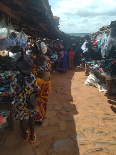 The image shows a bustling market in Lokoja, Nigeria. The coordinates (latitude: 7.404, longitude: 7.632) place the market on a dirt road, beneath a makeshift canopy of wooden beams and corrugated tin. The market is filled with vendors selling a variety of goods, including clothing, shoes, and bags. There are several people walking through the market, including two women, one carrying a small child on her back. The women are wearing brightly colored clothing.  The market is located in a densely populated urban area.  The surface of the road is made up of flat, irregular stones. The stalls and the surrounding environment are shaded by the bright sunshine.  The market is a vibrant place full of activity, with a strong sense of community.