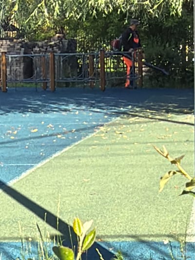A person is using a leaf blower on a playground in Shcherbinka, Russia. The person is wearing a black jacket with orange accents and orange pants. They are standing on a raised platform behind a metal fence. The playground surface is a combination of blue and green. Leaves are scattered across the playground. In the foreground, there is a shadow cast by a nearby object, and some green plants are visible. The background shows trees and a brick wall.