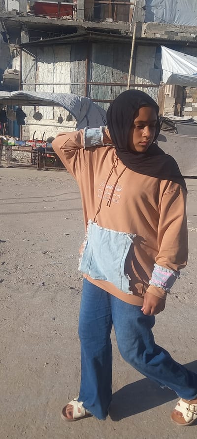 A young female wearing a black hijab, a light brown hoodie with distressed denim accents, and blue jeans stands on a dirt road in Khan Yunis, Palestinian Territories. The hoodie has a graphic design on the chest and distressed denim patches on the front pocket and sleeves. She has one hand on her head. Behind her, damaged buildings and makeshift structures are visible, indicating an urban environment impacted by conflict. A stall with goods is set up in the background. The ground is covered in loose dirt and gravel, and shadows suggest the presence of sunlight.