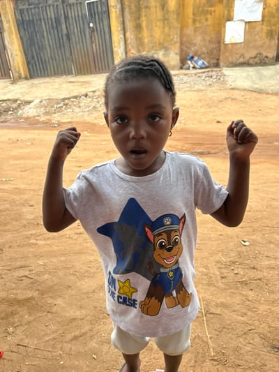 The image displays a young child positioned centrally outdoors in what is identified as the city of Ashipa, Nigeria. The child, of African descent, faces the camera directly, with an open-mouthed expression and wide eyes. Both arms are bent at the elbows, with fists clenched and raised to shoulder height. The child wears a light grey short-sleeved t-shirt featuring a large blue star and the character "Chase" from Paw Patrol, accompanied by the text "ON THE CASE" and a yellow star badge. Light-colored shorts are visible, and dark flip-flops are partially visible on the feet. The child has dark braided hair and wears small gold hoop earrings in both ears. The ground beneath the child is reddish-brown dirt. In the background, a faded yellow-ochre wall or building facade is visible. To the left, a dark, corrugated metal gate or door is present, with the number "49" faintly visible above and to its left. To the right on the yellow wall, two square white objects are affixed. A blue object, possibly a plastic bag, rests on the ground near the wall to the right. The scene is illuminated by bright, even daylight.