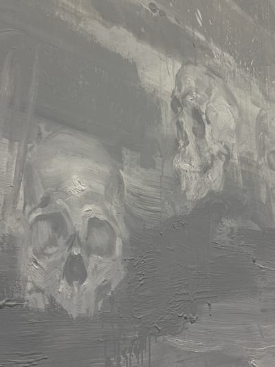 The image shows a close-up of a gray, textured surface, likely paint on a wall or canvas. The surface has visible brush strokes and layered application, creating variations in tone and depth. There are what appear to be vague, almost skull-like shapes formed within the texture, but they are not clearly defined. The lighting is soft and diffused. There are no people, subjects, objects, actions, or interactions. The environment is undefined due to the close-up view. The location in Abu Dhabi, United Arab Emirates, is not ascertainable from the image.