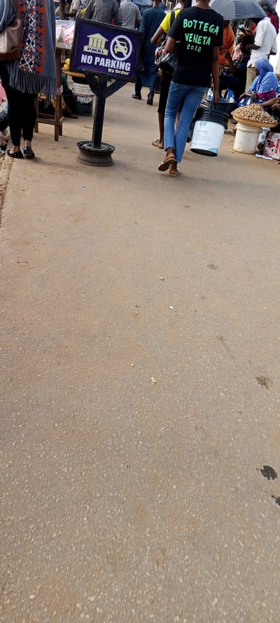 The image presents an outdoor street scene in Aso, Nigeria. The foreground consists of a paved surface with visible wear and small debris. A "NO PARKING By Order" sign is positioned on the left, mounted on a black post with a circular base. Several people are partially visible in the background, including one wearing a black "BOTTEGA VENETA 2022" t-shirt and blue jeans, carrying a white bucket. Other figures are seen walking or standing near makeshift stalls or displays, with one person beneath an umbrella. Commodities for sale, like peanuts, are apparent on a nearby table.