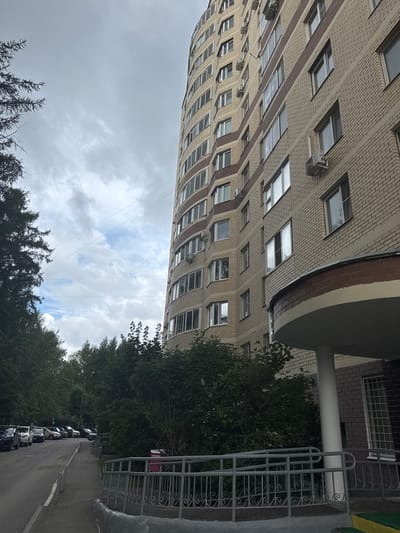 The image shows a view of a light-colored brick apartment building with a curved facade and numerous windows, some of which have air conditioning units installed. The building is in Zelenograd, Russia, as indicated by the context. To the left of the building, there is a street with parked cars and a sidewalk lined with trees and bushes. The sky is overcast with clouds. A metal railing is visible along the sidewalk in the foreground. The scene appears to be an ordinary residential street in a city.