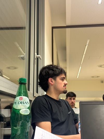 The image depicts an indoor scene within the city of Guimarães, Portugal, appearing to be a classroom or office environment.
In the foreground on the left, a green plastic bottle of "PEDRAS SALGADAS" carbonated mineral water, labeled "GÁS 100% NATURAL," stands upright with its green cap visible. The bottle partially obscures a window structure to its left.
A young adult male with dark, wavy hair, a short beard, and a goatee is seated in the mid-ground, facing right in profile. He is wearing a black short-sleeved t-shirt with a small white crocodile logo on the left chest. His right arm rests on a surface, beside which a sheet of white lined paper is visible.
Further back and to the right, a second young adult male with short dark hair and a mustache is partially visible, looking forward. He appears to have white earbuds in his ears. A black laptop screen stands in front of him, partially obscuring his body.
The background features light-colored walls and a white acoustical tile ceiling with a perforated pattern. The ceiling includes multiple long, rectangular fluorescent light fixtures and circular air vents. A large window with white framing is prominent on the left, exhibiting reflections of the interior ceiling and light sources. The lighting is artificial and consistent with an indoor institutional setting.