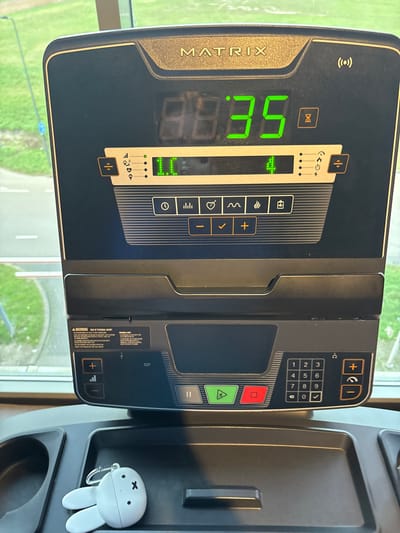 The image presents a close-up, eye-level view of a black and grey MATRIX brand treadmill console, positioned indoors overlooking an outdoor environment through a large window. The console's digital green LED display shows "8.8:35" with an hourglass icon to the right of "35." Below this, "II" and "4" are illuminated, accompanied by various icons representing metrics like incline, heart rate, calories, and power. The lower section of the console includes a "WARNING: RISK OF PERSONAL INJURY" label, a numeric keypad, and red, green, and grey buttons for stop, play, and pause functions respectively. On the flat tray section of the console, a white Miffy (Nijntje) character keychain or earbud case with black 'x' eyes and a horizontal line mouth rests. Through the window, a paved road with white markings is visible, curving to the left, bordered by green grass. A blue pole is partially observable on the left side of the outdoor scene. The setting is within a fitness center in Rosmalen, Netherlands.