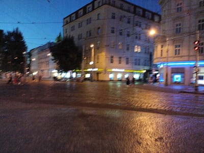 The image depicts a street scene at dusk or night in Prague, Czechia. The street is paved with cobblestones, and the surfaces are wet, reflecting the artificial lights. Buildings line the street, with illuminated windows and storefronts. Some of these storefronts have bright blue and yellow lighting. A street lamp is visible, casting a warm glow on the scene. Traffic lights are also present, with a red light illuminated. Trees are visible on the left side of the image, silhouetted against the twilight sky. The sky is a deep blue, indicating the late hour. There are some blurry figures of people walking on the sidewalk and crossing the street. The overall impression is of a bustling urban environment at night, possibly after a rain shower, with the lights creating a vibrant atmosphere. The motion blur suggests that the photo was taken while the camera was moving, or with a slow shutter speed.