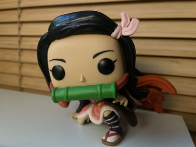 A close-up, eye-level shot focuses on a Nezuko Kamado Funko Pop figure, a character from the anime series Demon Slayer, positioned on a white surface. The figure is in a crouching pose, with its signature bamboo muzzle in place. Behind the figure, a set of horizontal blinds creates a textured backdrop, illuminated by a warm, ambient light source, suggesting an indoor setting. The figure's details, including its black hair adorned with a pink hair accessory, large eyes, and the patterned leggings and sandals, are clearly visible. The overall scene is static and well-lit, emphasizing the collectible figure. There is no indication of the image being taken in Warsaw, Poland, or any specific city.